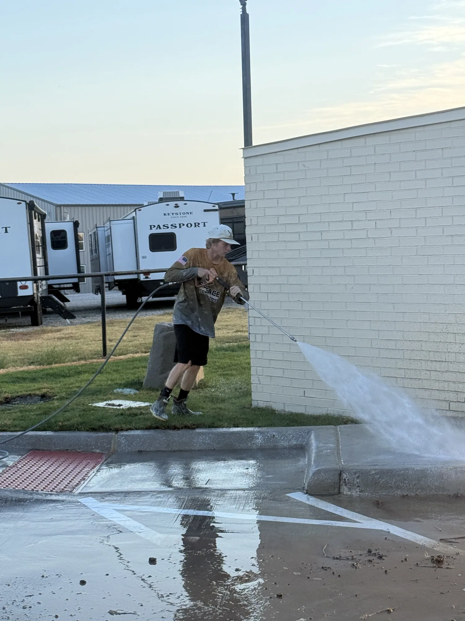 Crew member in a company shirt pressure washing the white brick wall and concrete apron of a commercial building, with RV trailers parked behind.